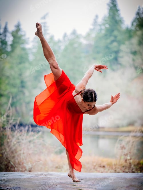 Preview: Dancer in a vibrant red dress performs a graceful ballet pose by a serene forest lake. USA