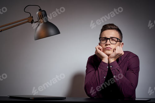 Preview: business man with glasses and a shirt sitting at a table in a dark room with a lamp cropped view
