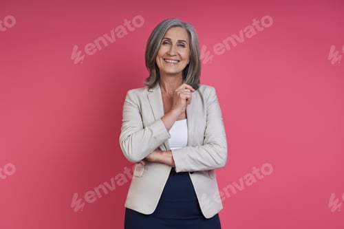 Preview: Confident senior woman in formalwear smiling while standing against pink background