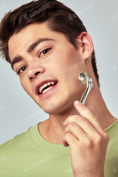 Preview: A young handsome smiling guy with brown hair uses a massage roller on his face. Caucasian man in the
