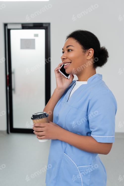 Preview: cheerful multiracial healthcare worker in blue uniform holding paper cup with takeaway coffee and