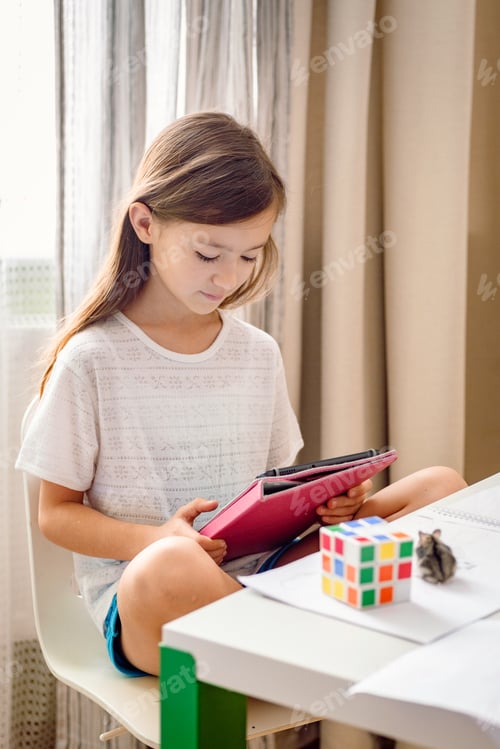 Preview: Caucasian school age girl sitting at the messed desk using tablet. Generation Z girl playing game.