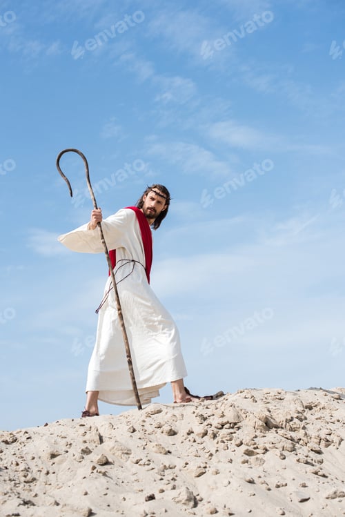 Preview: low angle view of Jesus in robe, red sash and crown of thorns walking on sandy hill with staff in