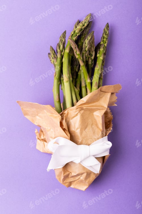 Preview: Directly above view of asparagus bunch in brown paper tied with white ribbon on purple background