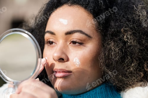 Preview: Head-shot of chubby young woman applying cream on face with mirror in front.