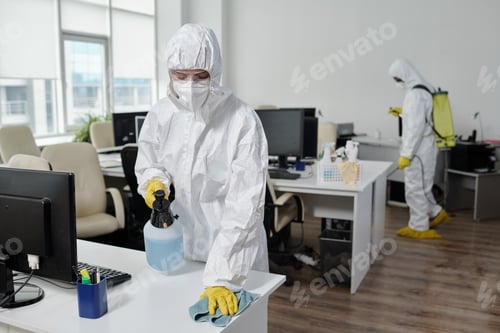 Preview: Young female in hazmat suit wiping desks while spraying disinfectant on surface against her male