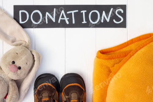 Preview: Children's plaid, plush toy hare and boots with a chalk Donations sign on a white wooden table. Flat