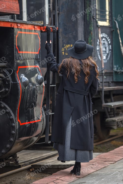 Preview: A beautiful girl in a black coat and hat with a suitcase in her hands near an old steam locomotive.