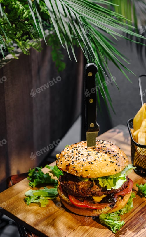 Preview: Burger with knife on wooden board next to fries in outdoor setting
