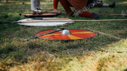 Preview: Medieval weapons, a red round shield and a steel sword lying on the battlefield in the open air.