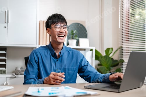 Preview: A smiling man is sitting at a desk with a laptop and smiling. He is wearing