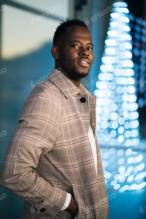 Preview: Elegant black businessman posing with hand in pocket near christmas lights