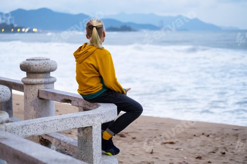 Preview: Lonely teenager looking to sea sitting on the waterfront. Rear view of teenager against seascape.