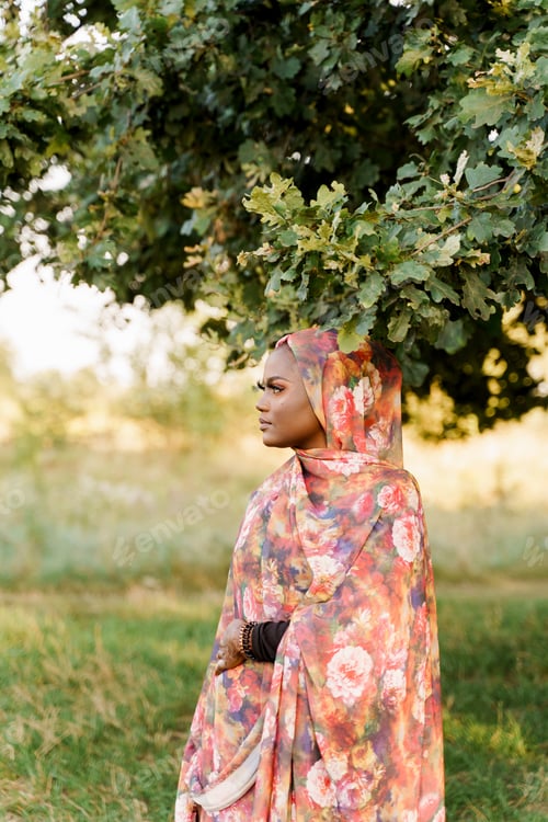 Preview: Black Muslim woman in colorful hijab smiling, looking left under green tree, calm moment