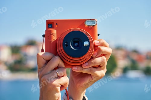 Preview: Hand holding instant camera photographing seascape with coastal town
