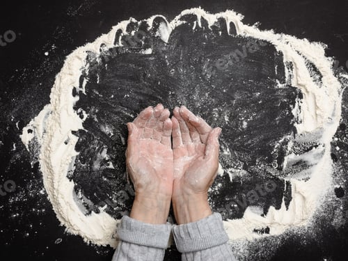 Preview: Sifted white wheat flour on a black table and two female hands, top view. cooking at home