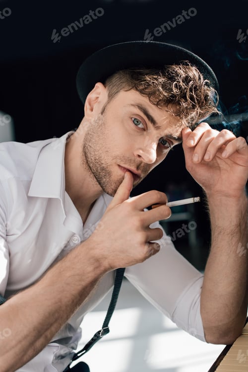 Preview: pensive handsome man in white shirt and black hat smoking cigarette