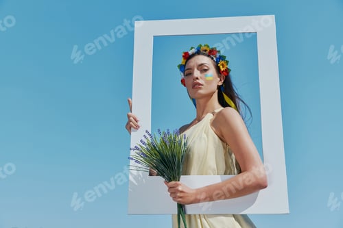 Preview: Attractive Ukrainian woman in floral crown holding picture frame with blue sky in the background