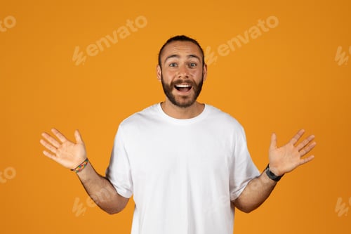 Preview: Smiling surprised caucasian millennial man with beard in white t-shirt spreads arms to sides