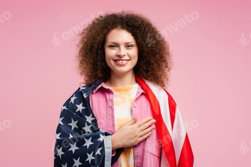 Preview: Attractive, smiling, happy young woman, patriot holding American flag with hand on heart