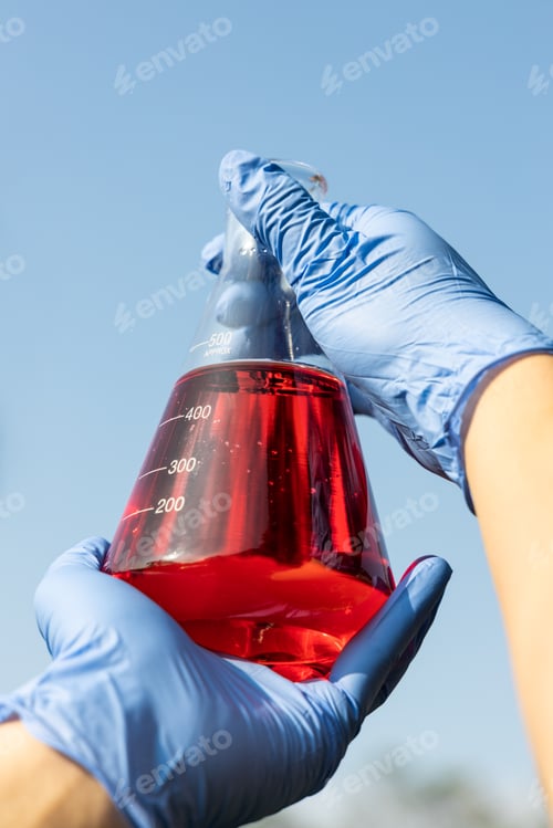 Preview: Hand with gloves holding beaker with red color unrefined palm oil against blue sky