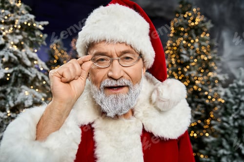 Preview: Santa Claus with glasses smiles in a snowy forest adorned with twinkling holiday lights