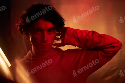Preview: Young Man with Wavy Hair Wearing Hoodie Standing in Red and Orange Light