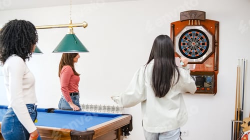 Preview: Woman throwing arrow at dartboard while female friends standing at pool table in game room
