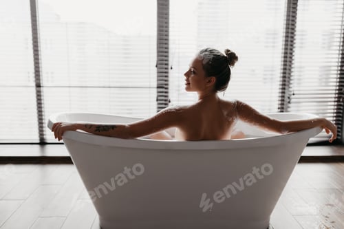 Preview: Back view of young lady taking foamy bath, looking aside at free space at home or hotel bathroom