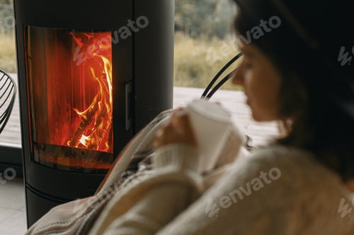 Preview: Fire burning in modern black fireplace and blurred image of woman in sweater holding cup of tea