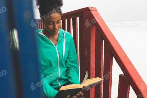 Preview: Front view of beautiful young Mixed-race woman reading book while sitting at beach hut