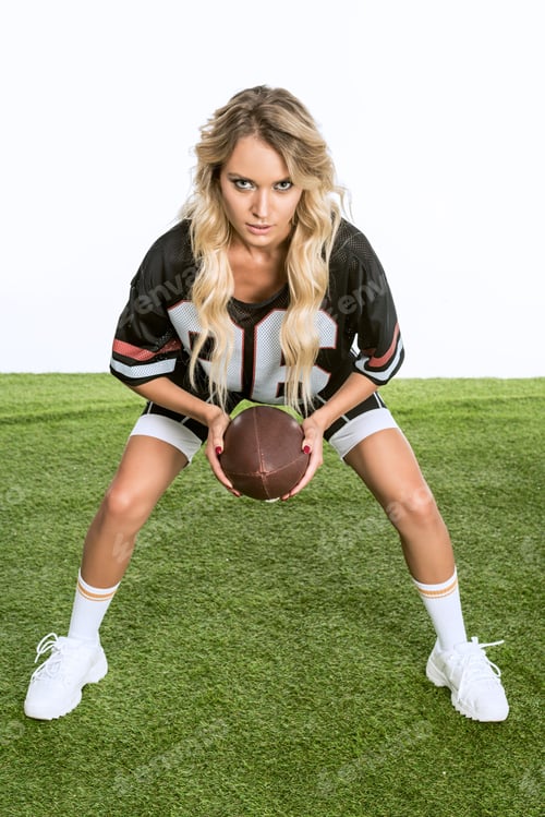 Preview: serious young woman in american football uniform holding ball while standing on green grass isolated