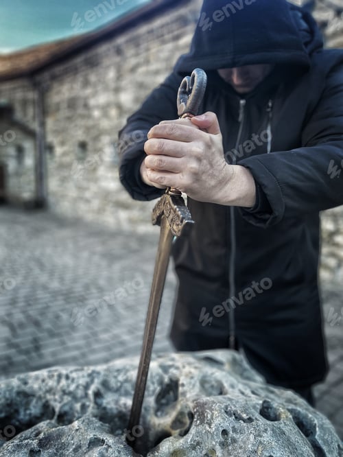 Preview: the sword in the stone of King Arthur - Clarent. a man in black drags an iron sword from a stone