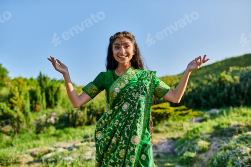 Preview: cheerful young indian woman in green sari posing and looking at camera on summer field