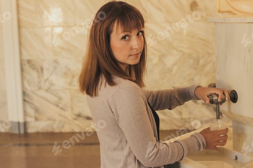 Preview: young girl with brown hair in a beige sweater pours mineral water into a glass in a drinking gallery
