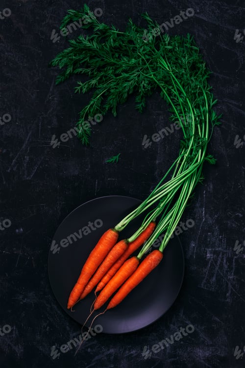 Preview: Ripe carrot with greens on black plate on black background. Healthy vegan food. Top view, flat lay
