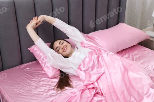Preview: Smiling woman stretching in bed with pink silk linens indoors