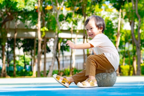 Preview: A happy young Asian boy sitting on a soccer ball at a bright playground in a park.