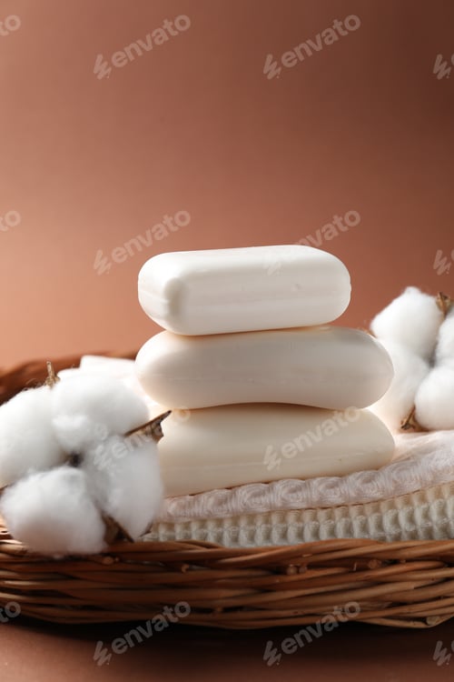 Preview: White soap bars and cotton flowers in wicker basket on brown background, closeup