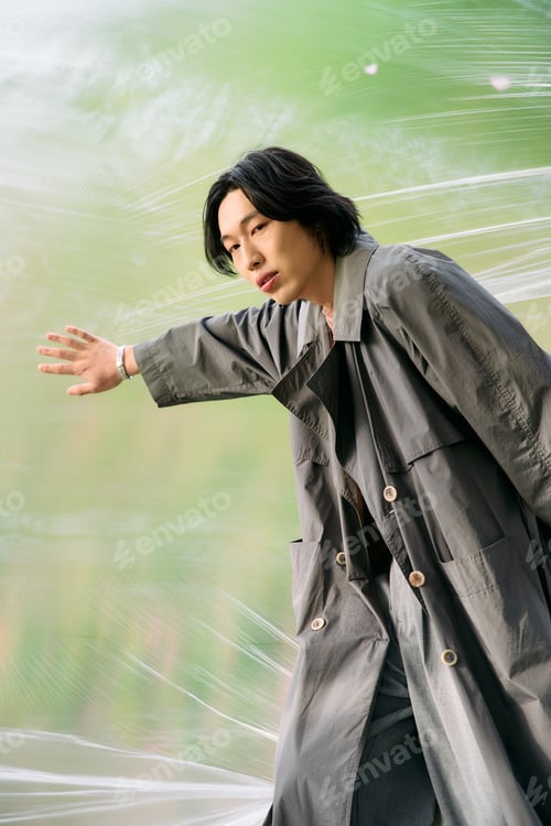 Preview: Stylish young man exploring nature in a grey trench coat during a serene park visit