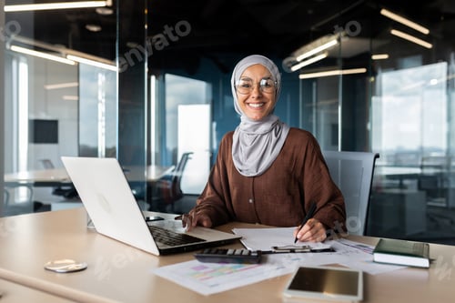 Preview: Portrait of young successful Arab woman in hijab working in office with documents and laptop
