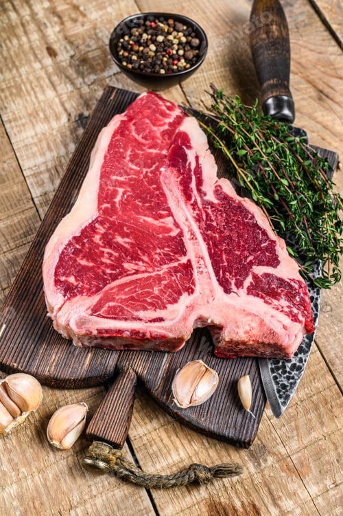 Preview: Raw Porterhouse or T-bone beef meat Steak with herbs on a wooden cutting board.