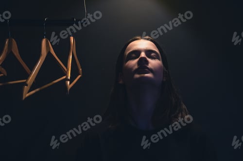 Preview: A young woman near empty hangers in the dark under a lamp.