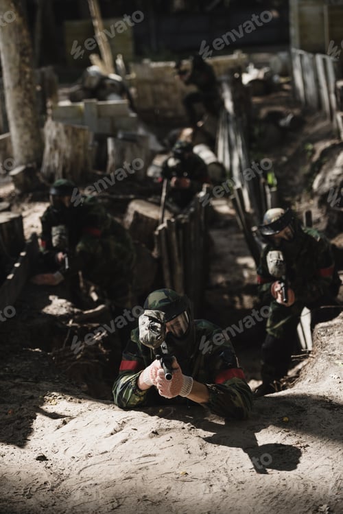 Preview: paintball team in camouflage and protective masks aiming by marker guns from ditch outdoors