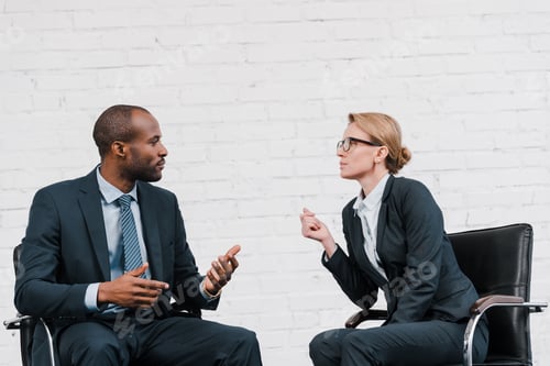 Preview: side view of african american businessman gesturing near businesswoman in glasses sitting on chair