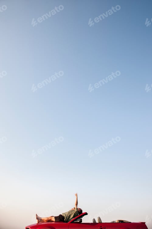 Preview: Caucasian male relaxing on the hood of his convertible sports car.