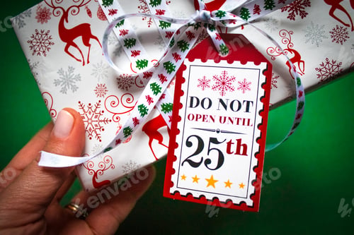 Preview: on a green background a gift box in hands with a tag Do not open until December 25