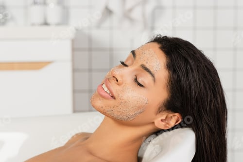 Preview: african american woman with closed eyes and scrub on face lying in bathtub
