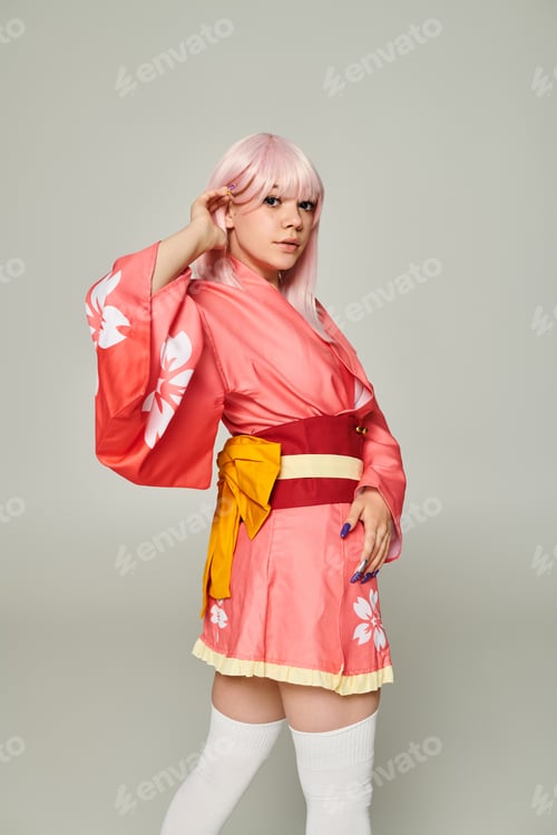 Preview: charming woman in colorful japanese style attire adjusting blonde hair and looking at camera on grey