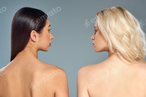 Preview: multiethnic models with bare shoulders looking at each other on blue backdrop, diverse beauty